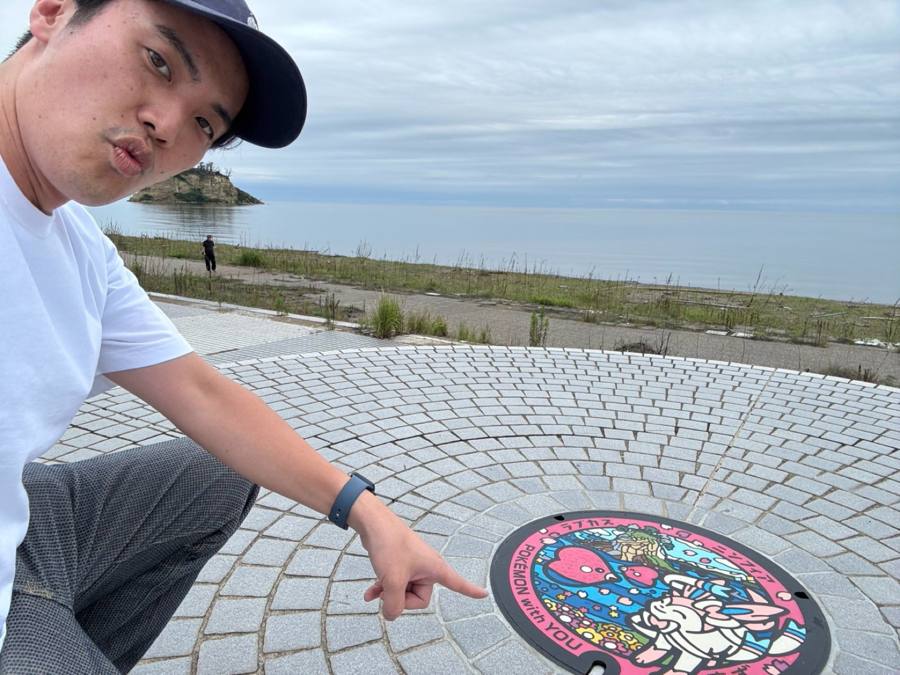 マンホールを指し海を背にする男性