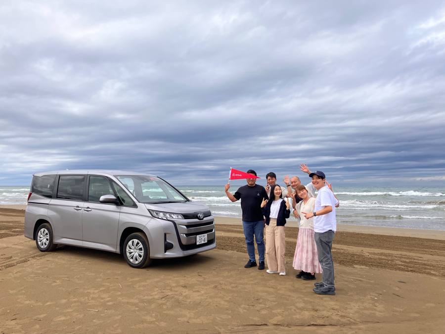浜辺で車と記念撮影、フラッグ持つ